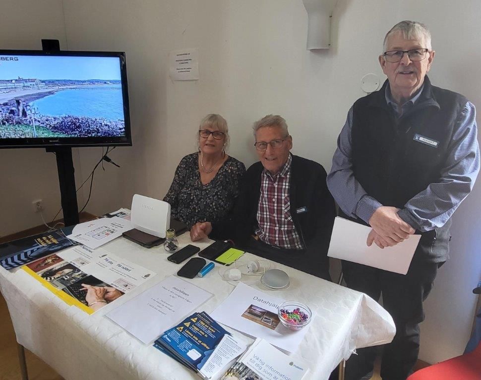 Två personer sitter och en person står bakom ett bord. Vid sidan av bordet finns en bildskärm.