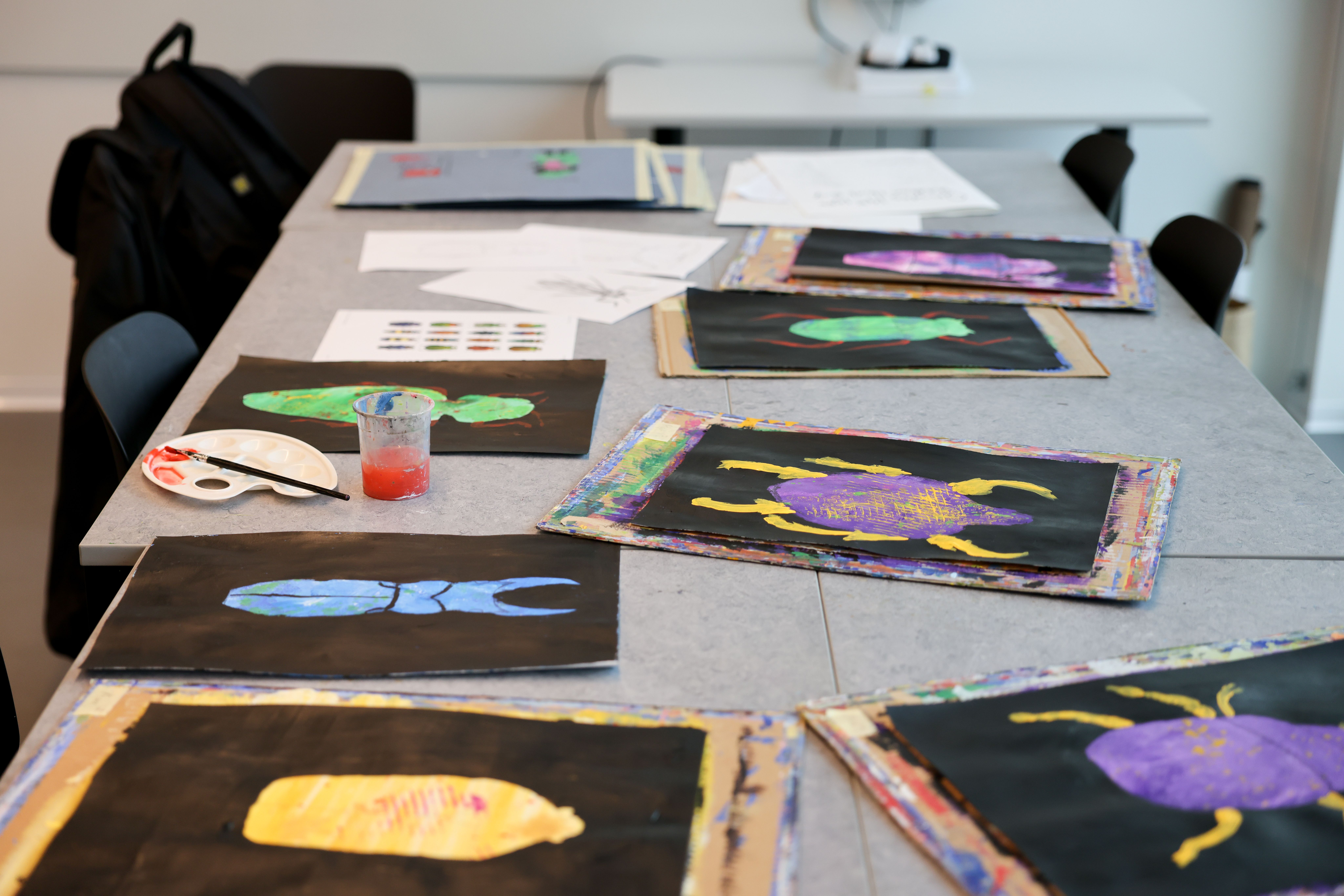 Målningar föreställande blåa, lila, gröna och gula skalbaggar ligger på ett bord.