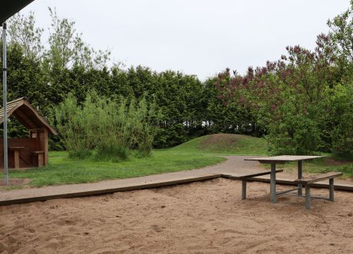 Sandlåda med bord och bänkar, lekstuga och kulle i bakgrunden
