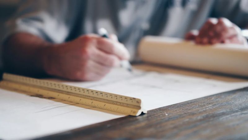Händer med penna och linjal vid en ritning på ett bord.