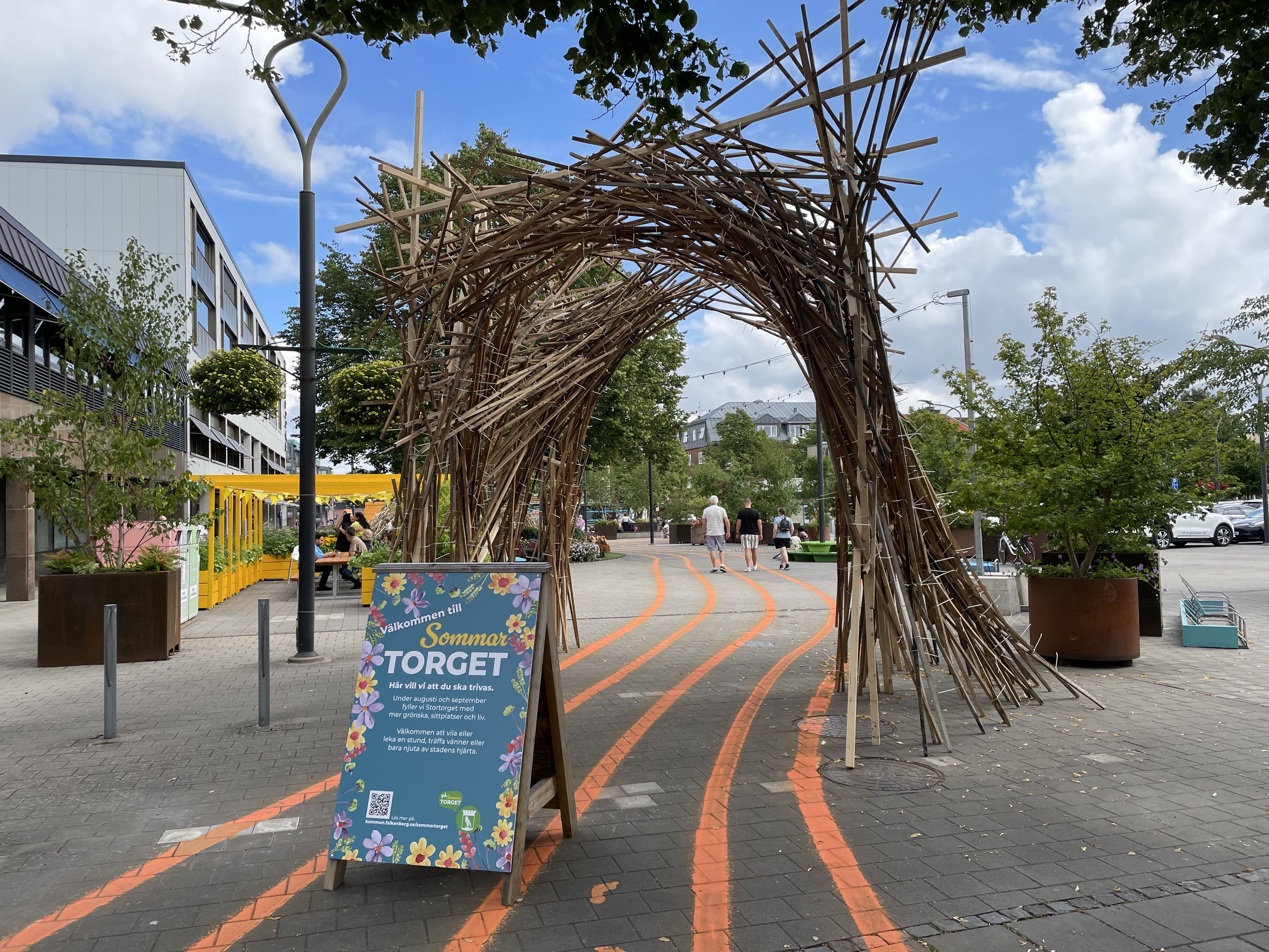 Konstverk av trä i en båge, linjer som du ska följa på marken och välkomstskylt vid entrén till sommartorget.