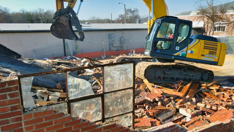 Grävmaskin på nerrivet hus med söndring tegelsten och söndrigt fönster.
