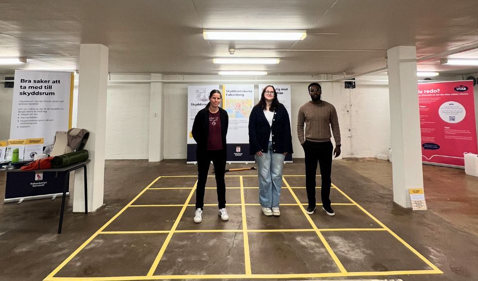 En bild från ett skyddsrum. Tre personer som står uppställda i ett rutnät, som illusterar hur mycket plats varje person beräknas ha i ett skyddsrum 0,75 kvadratmeter.