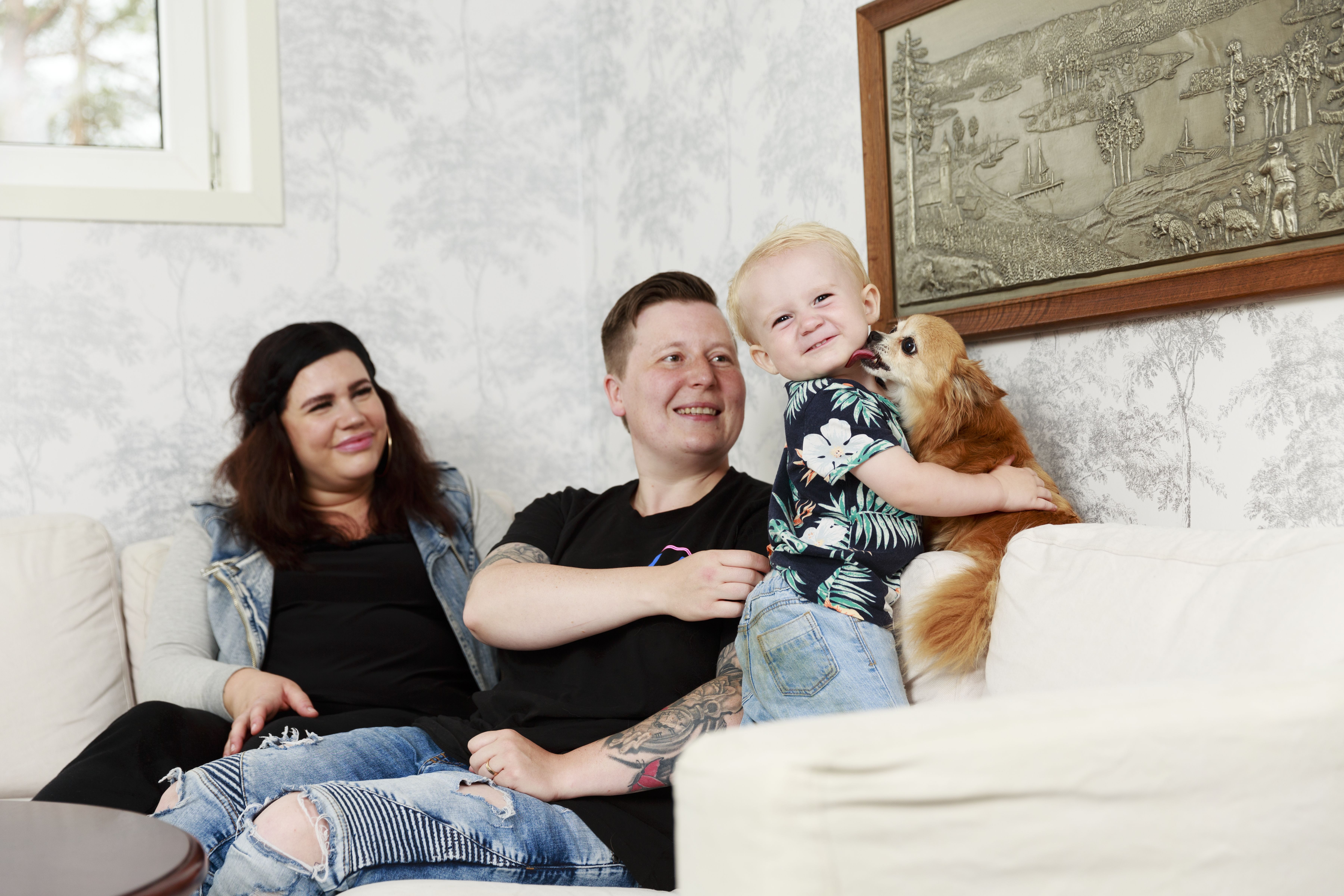 Två kvinnor sitter i en soffa. Framför dem står en liten pojke som håller om en hund.