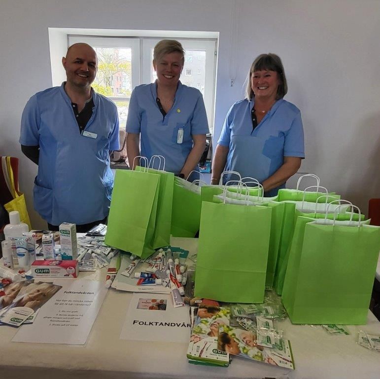 En man och två kvinnor i blå kläder står bakom ett bord. På bordet står gröna papperspåsar.