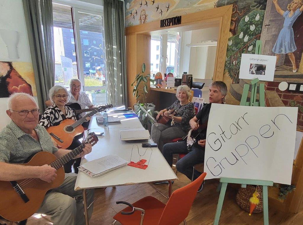 Äldre personer med varsin gitarr i knäet sitter runt ett bord. På en skylt vid sidan av bordet står det Gitarrgruppen.