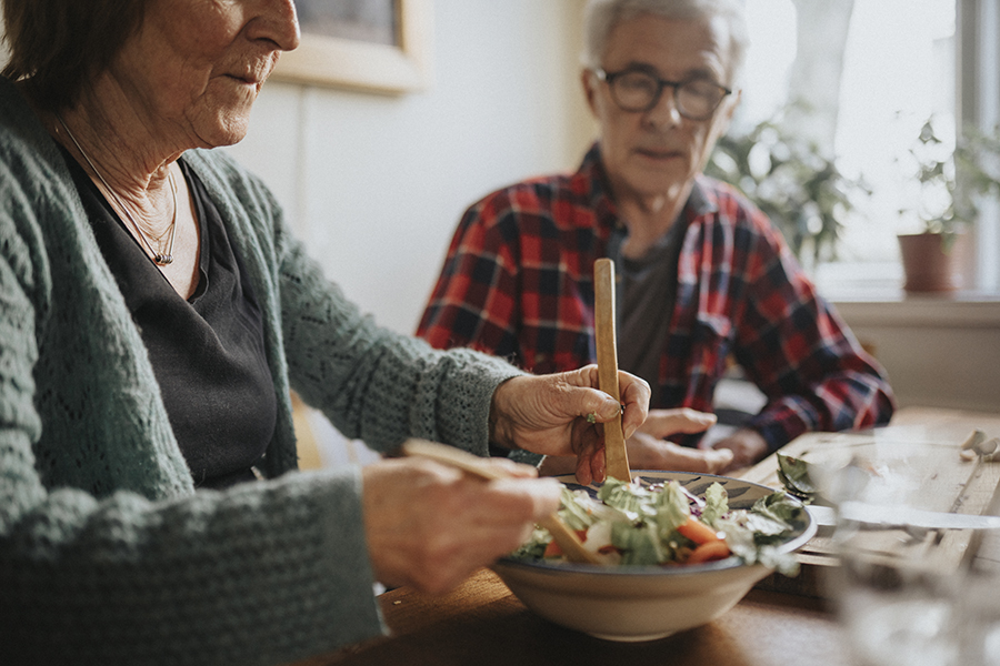 Ett äldre par sitter vid ett köksbord. Kvinnan tar sallad ur en skål.