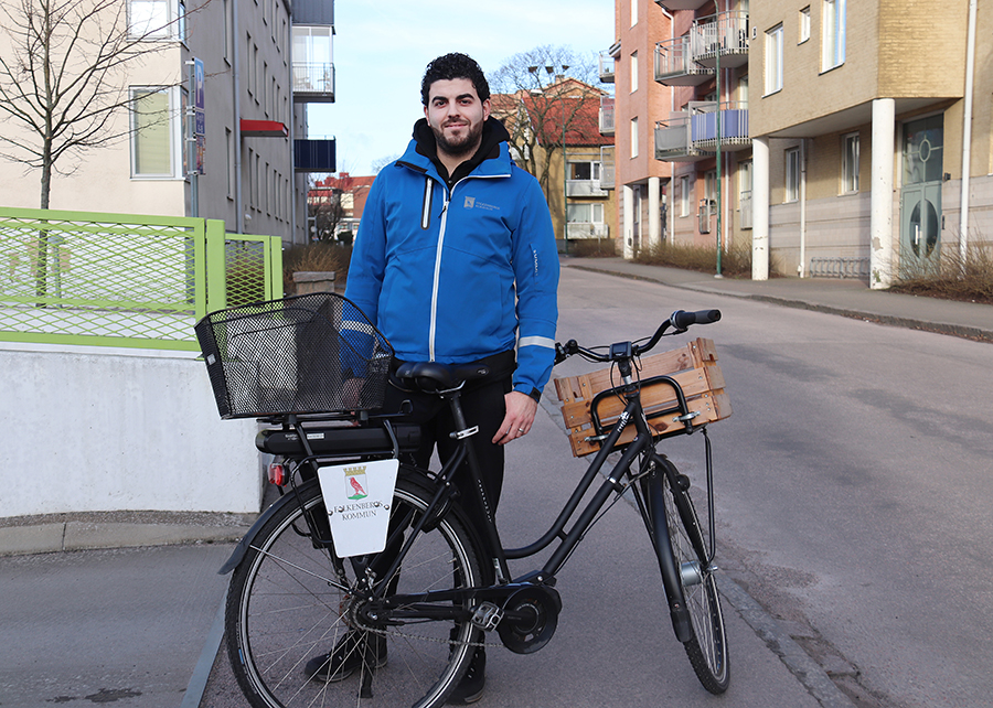 En man med mörkt hår och blå jacka står ute på en trottoar, bakom en svart cykel.