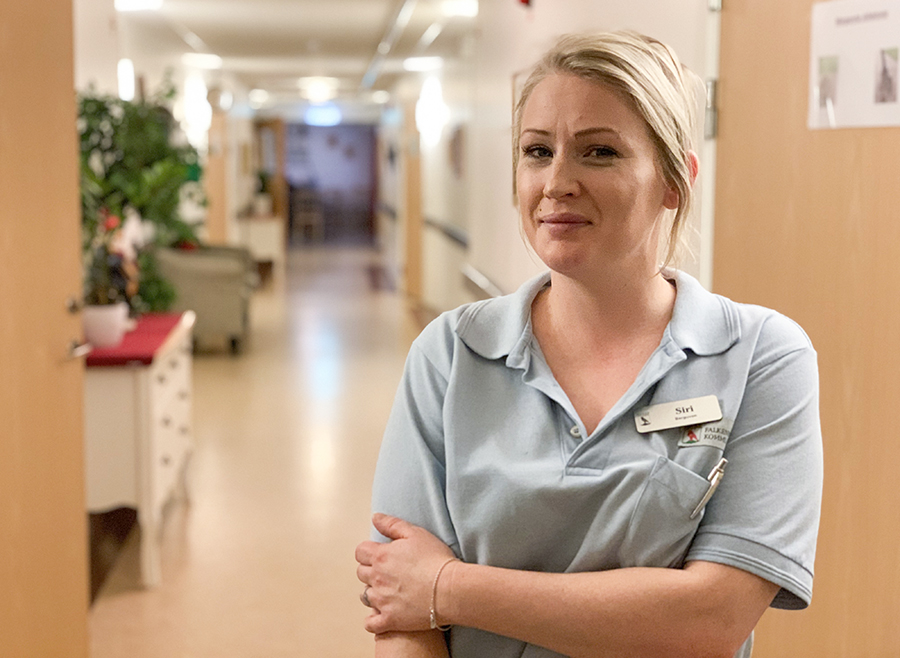 Leende, blond tjej med ljusblå t-shirt står i korridoren på ett särskilt boende för äldre.