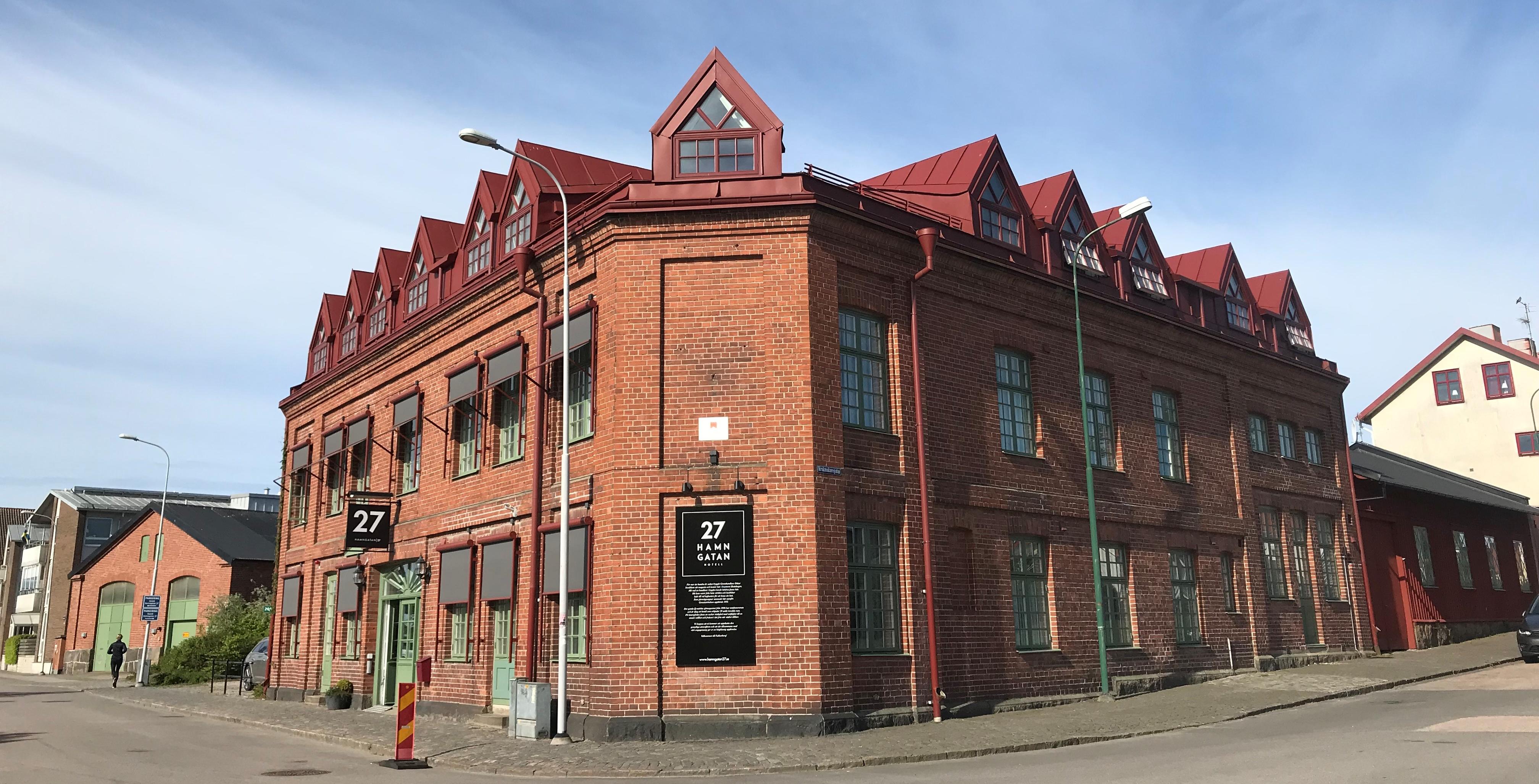 Hotell Hamngatan 27 är en röd tegelbyggnad med gröna fönster och röd takkupor. Det är blå himmel med skira moln.