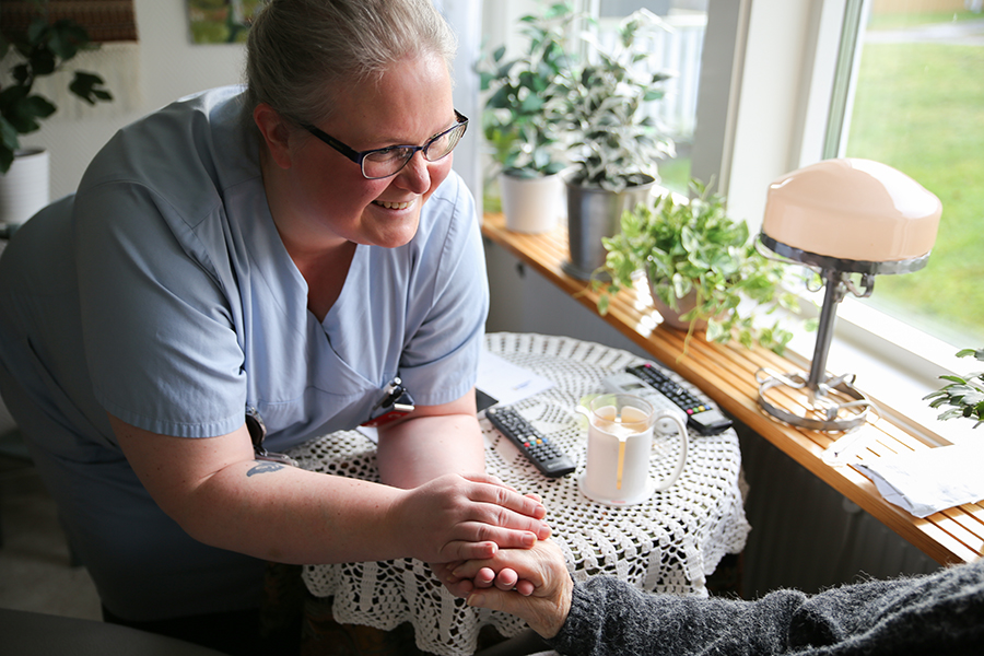 En leende kvinnlig undersköterska lutar sig fram över ett bord och håller en äldre person i handen.