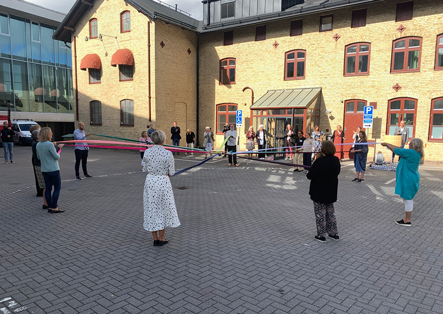 Elva personer står i en cirkel och håller färgglada band, som knutits ihop i mitten, mellan sig.