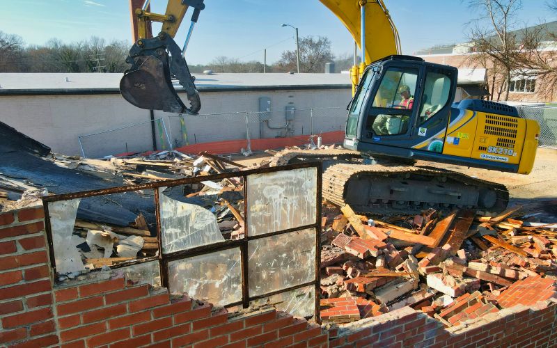 Grävmaskin på nerrivet hus med söndring tegelsten och söndrigt fönster.