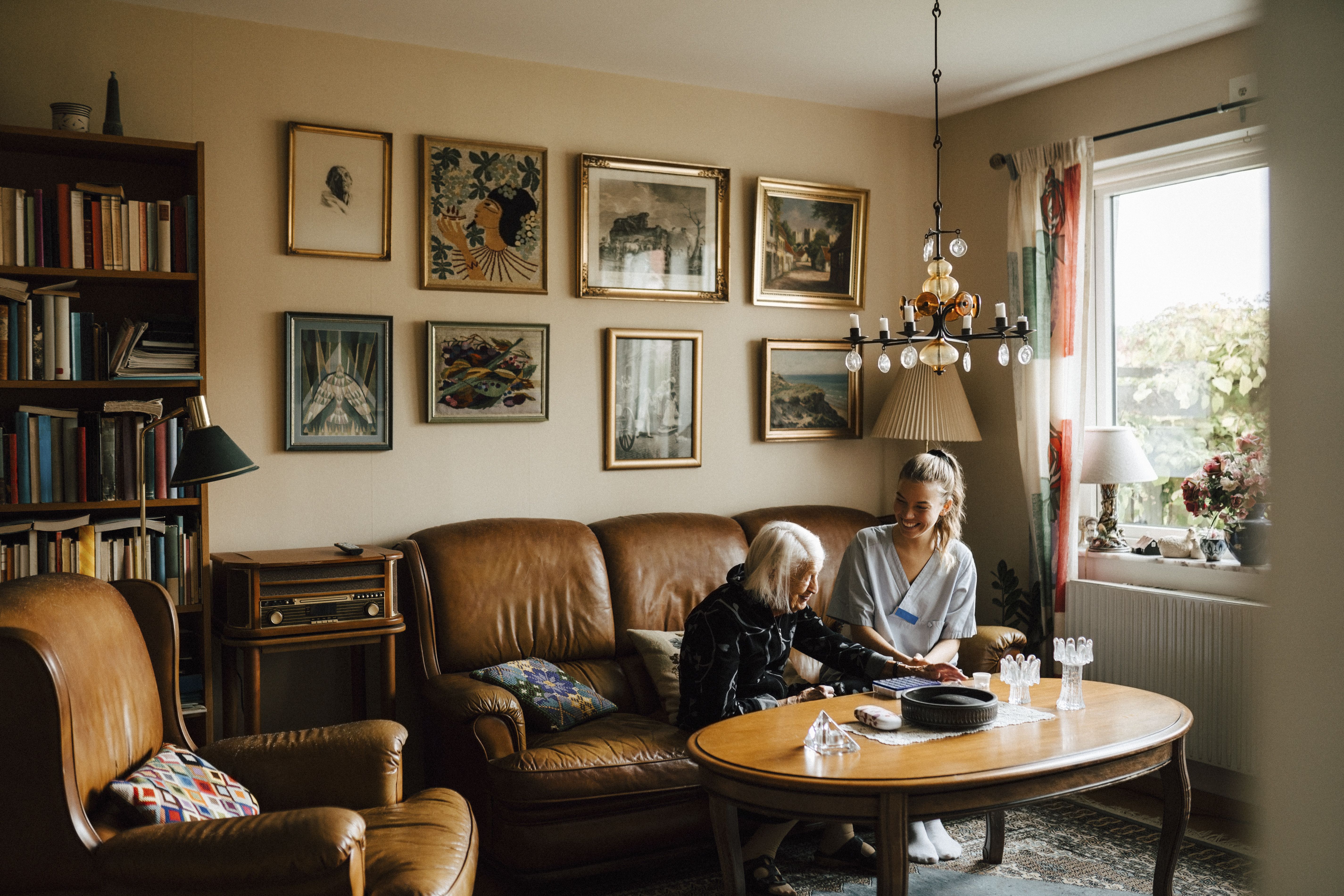 Kvinnlig undersköterska pratar med äldre kvinna på en soffa i ett vardagsrum.