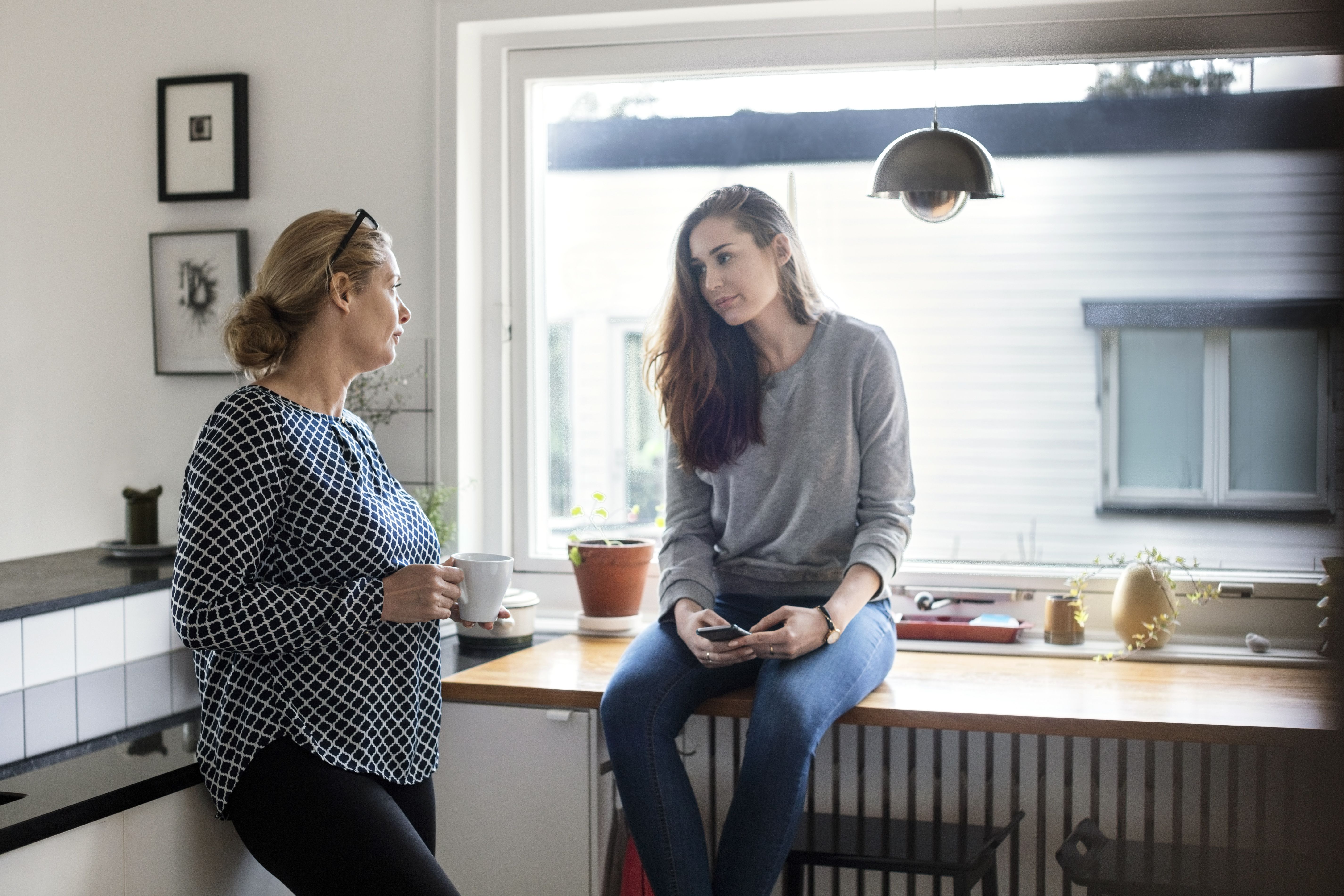 Två kvinnor diskuterar något i ett kök, en med an kopp i handen, en annan sittandes på köksbänken med en mobil i handen.