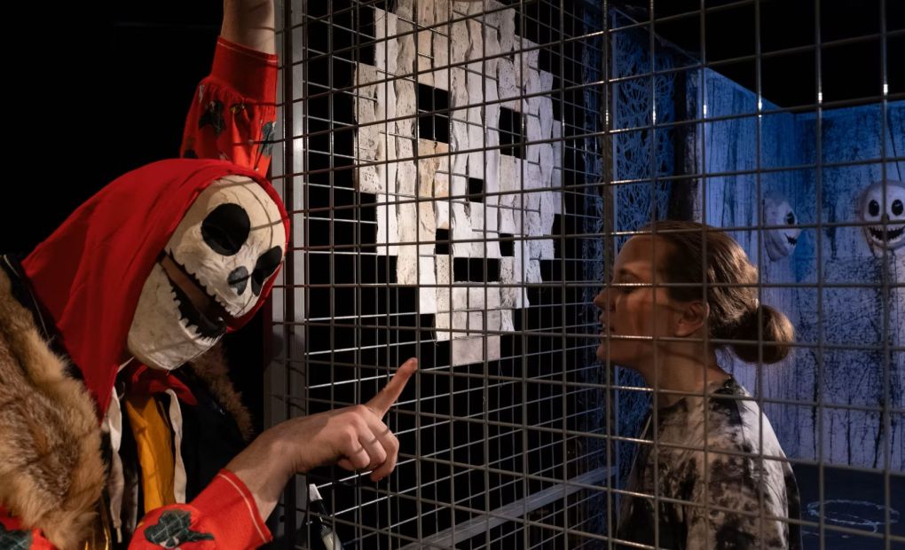 Bild från föreställningen Galten. En person i en gallerbur och en maskerad rödklädd person står utanför och pekar in.