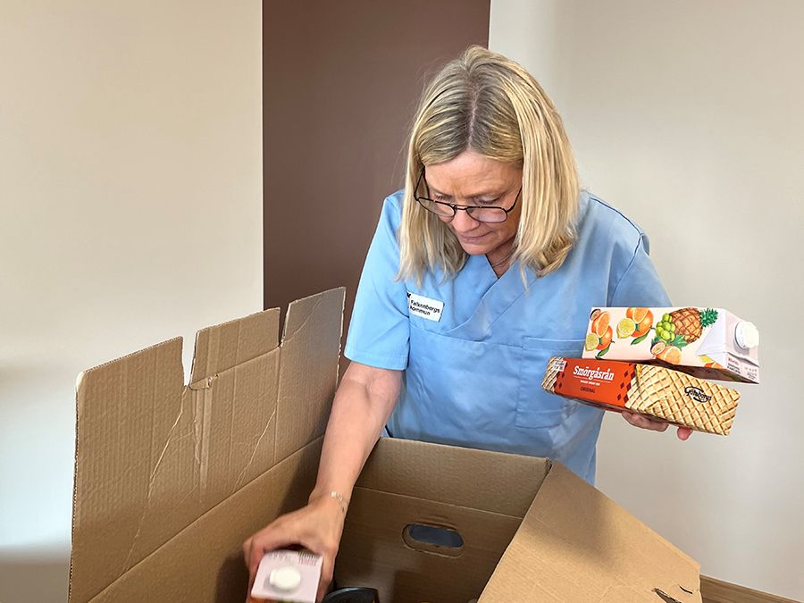 Blond kvinna med blå arbetströja packar upp en låda med matvaror.