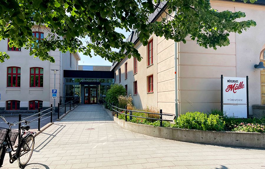 En gång leder in till en entré mellan två beigefärgade stenhus. Framför det högra huset står det en skylt där det står Mötesplas Mölle.