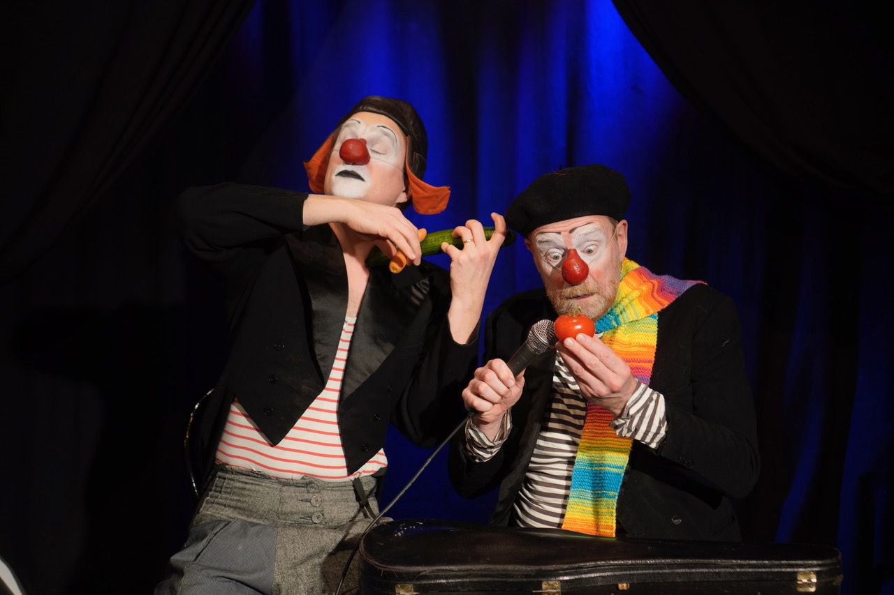 Två män sminkade som clowner med röda näsor, färgglada tröjor och frack. Den ena clownen håller i en tomat, den andra håller i en gurka.