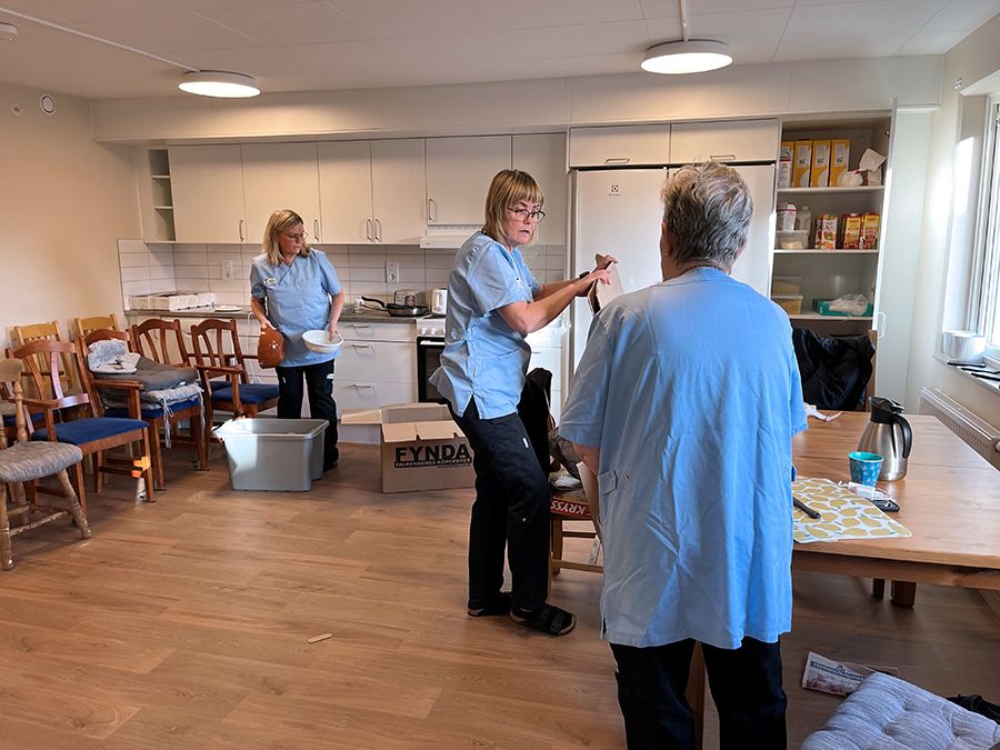 Tre kvinnor i blå arbetströjor packar upp saker i ett kök.