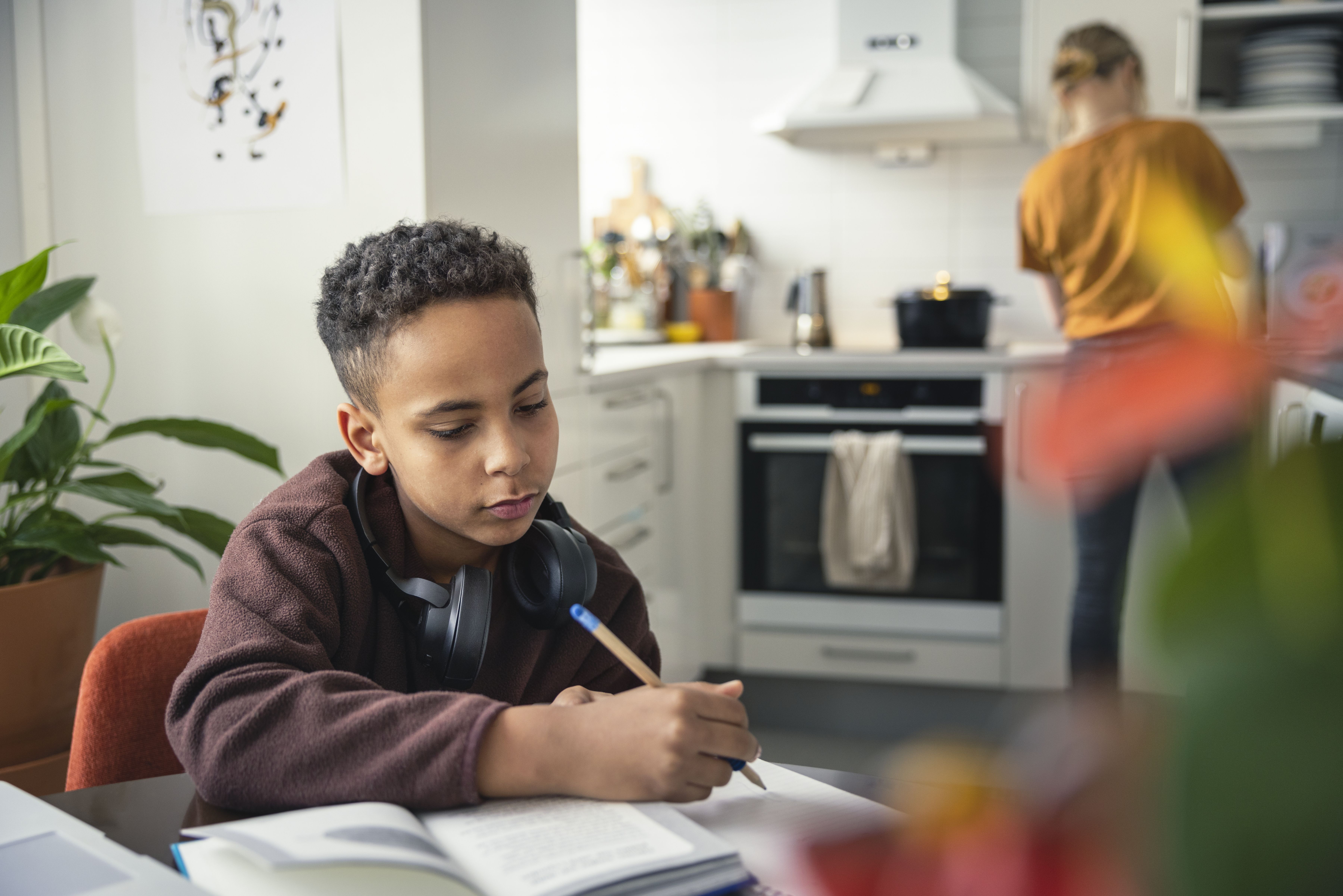En ung mörkhyad kille sitter vid ett köksbord med skolböcker framför sig. I bakgrunden står en ljushårig kvinna och lagar mat.