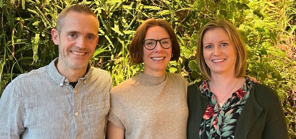 Personerna ståt framför en vägg med växter. Det är en överkroppsbild. De ler.