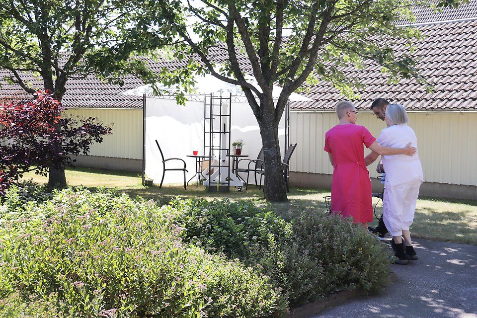 Två undersköterskor går bredvid en äldre dam ute i en trädgård. 