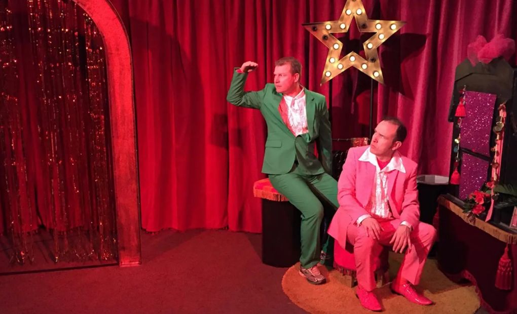 Två män i färgstarka kostymer sitter på en scen med rött tyg i bakgrunden. Från föreställningen Jag sa ju förlåt.