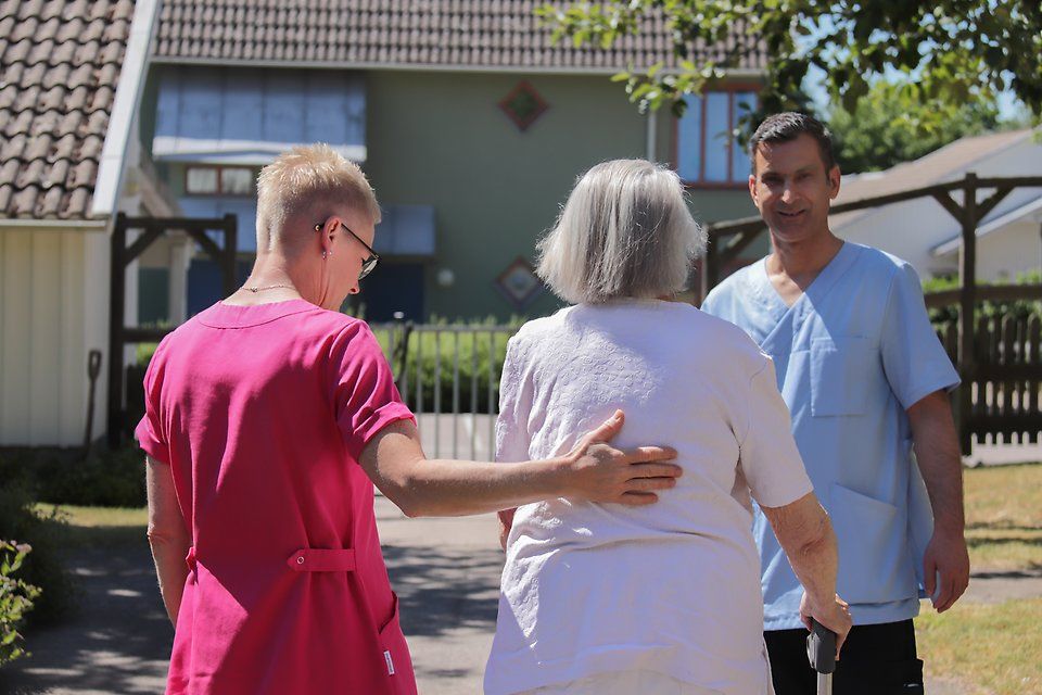 En kvinnlig undersköterska med kort hår går utomhus bredvid en äldre dam och håller sin hand på hennes rygg. En manlig undersköterska syns i bakgrunden.