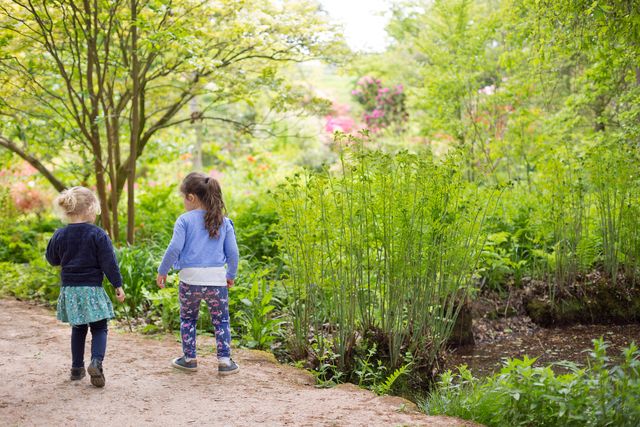 Två små flickor, 3-4 år, håller hand och går i en trädgård eller park.