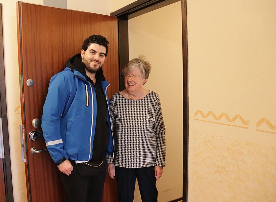 En man med mörkt hår och blå jacka står bredvid en äldre dam i dörröppningen till en lägenhet.