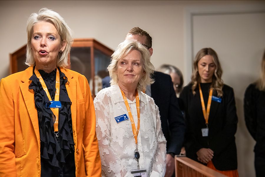 Blond kvinna i orange kavaj pratar. Till höger om henne står en blond kvinna i vit blus.