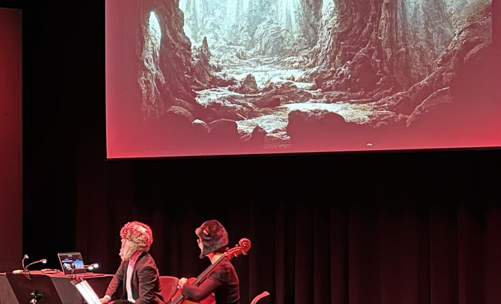 Två personer med instrument sitter på en scen , i bakgrunden en bild av en grotta med texten "60 000 år före vår tideräkning"