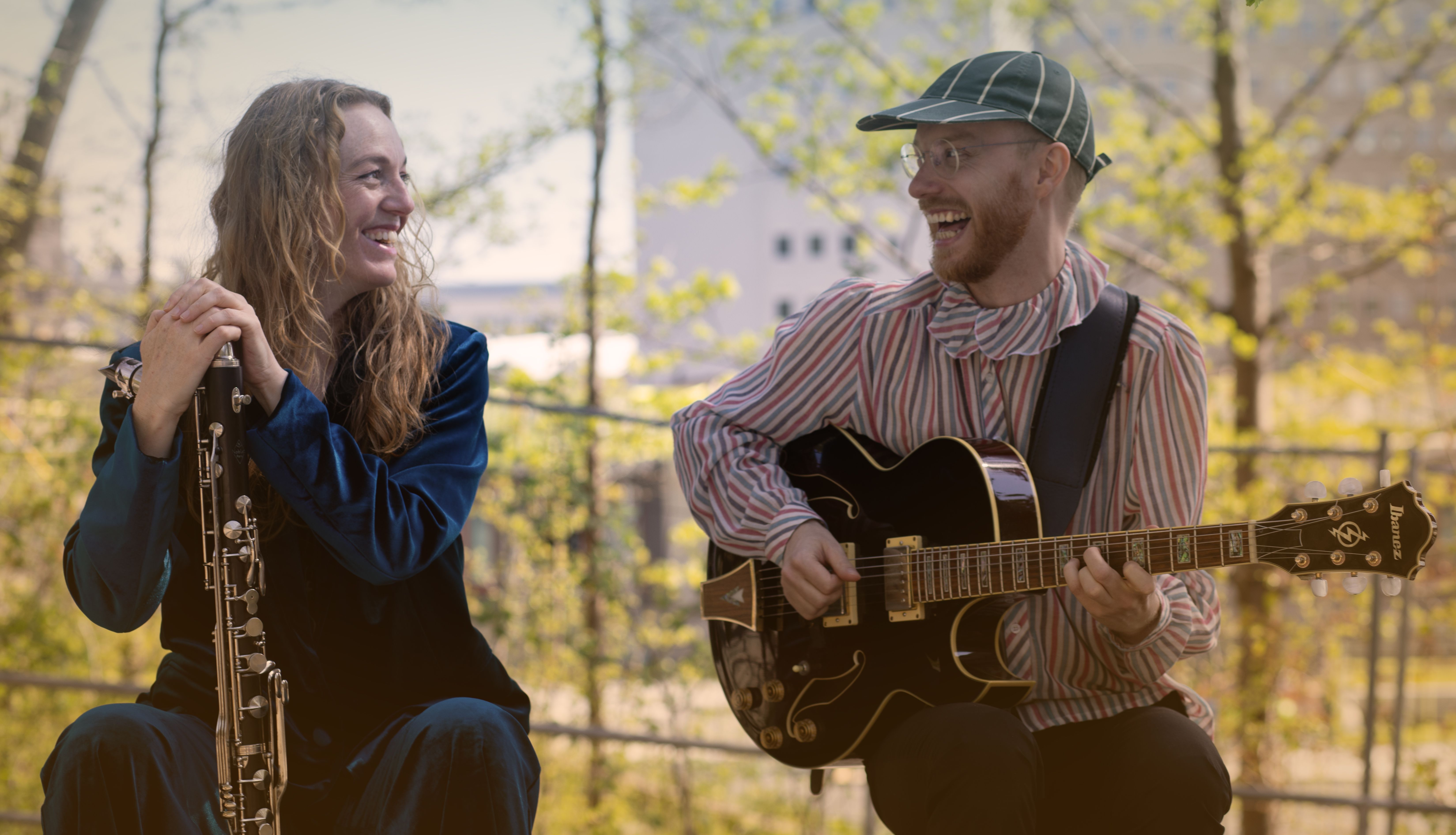 En kvinna och en man sitter bredvid varandra med träd i bakgrunden. Mannen spelar på en gitarr och kvinnan håller i en basklarinett.